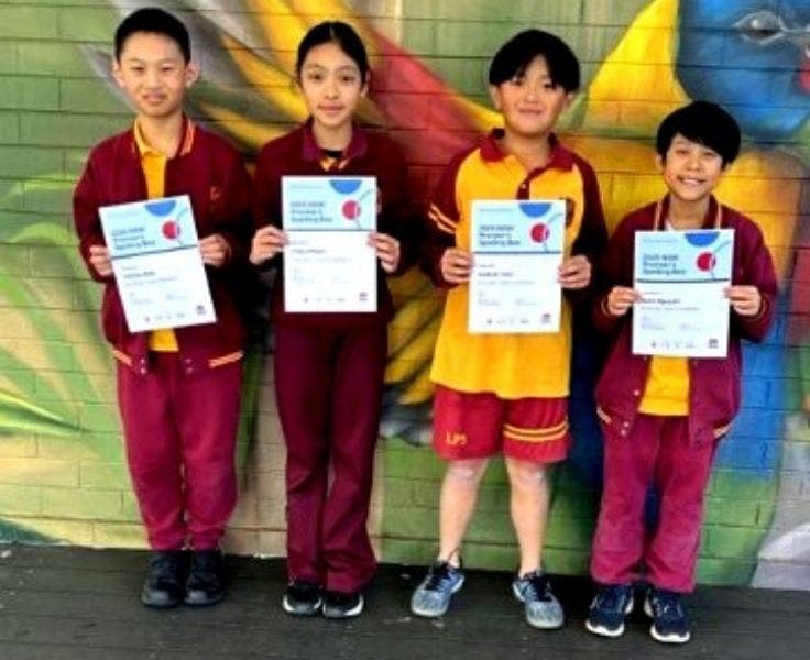 Four students in maroon and yellow uniforms are standing in front of a colorful mural, proudly holding certificates of achievement. The vibrant background adds to the celebratory atmosphere as they display their awards.