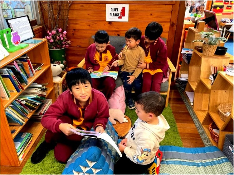 Several children are gathered in a cozy reading corner of a library or classroom, surrounded by shelves of books. Some are dressed in maroon and yellow uniforms, and they are reading storybooks together on a green rug and a couch. The atmosphere is warm and inviting, with colorful decorations, flowers, and a