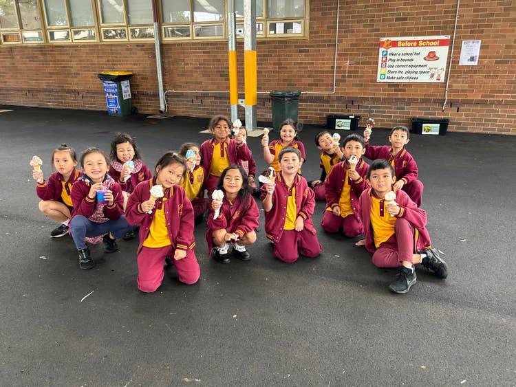 A group of students in maroon and yellow uniforms are sitting together on an outdoor paved area, smiling and holding drinks, with a school building in the background. The scene welcomes visitors to Lansvale Public School, highlighting a friendly and inclusive school community.