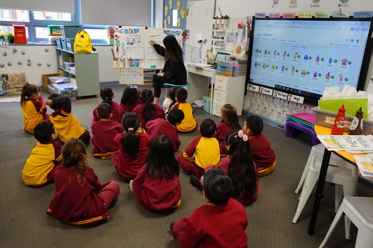 Young students in maroon and yellow uniforms sit on the classroom floor, attentively listening to their teacher, while a digital interactive whiteboard is used for engaging lessons, highlighting growth in the classroom and beyond.