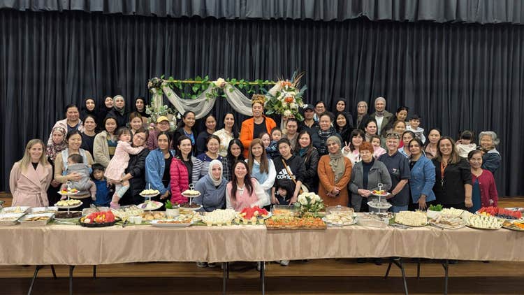 A large group of parents, staff, and children are gathered together in front of a beautifully decorated table filled with a variety of food and treats. They are posing in a hall with a backdrop of dark curtains and floral arrangements, celebrating the Parents and Citizens Association (P&C) and highlighting the importance of school community involvement and support.